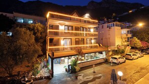 A multi-story hotel with balconies, a ground floor restaurant, and cars parked in front. - İsinda Pansiyon (Kas)