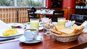 A breakfast setting with plates of scrambled eggs, toast, and a bowl of cereal. There are cups of coffee and juice on the table.