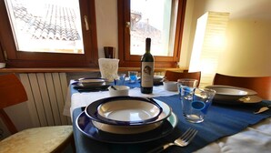 Une table à manger dressée avec des assiettes, des couverts, des verres et une bouteille de vin, avec vue sur un toit recouvert de tuiles, visible à travers une fenêtre.