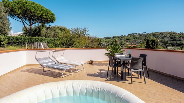 An outdoor hot tub with lounge chairs and a table, surrounded by a wooden deck and a view of a hill.