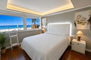 Un dormitorio con una cama grande, una ventana con vistas a la playa, una mesita de noche con lámpara y una obra de arte colgada en la pared.