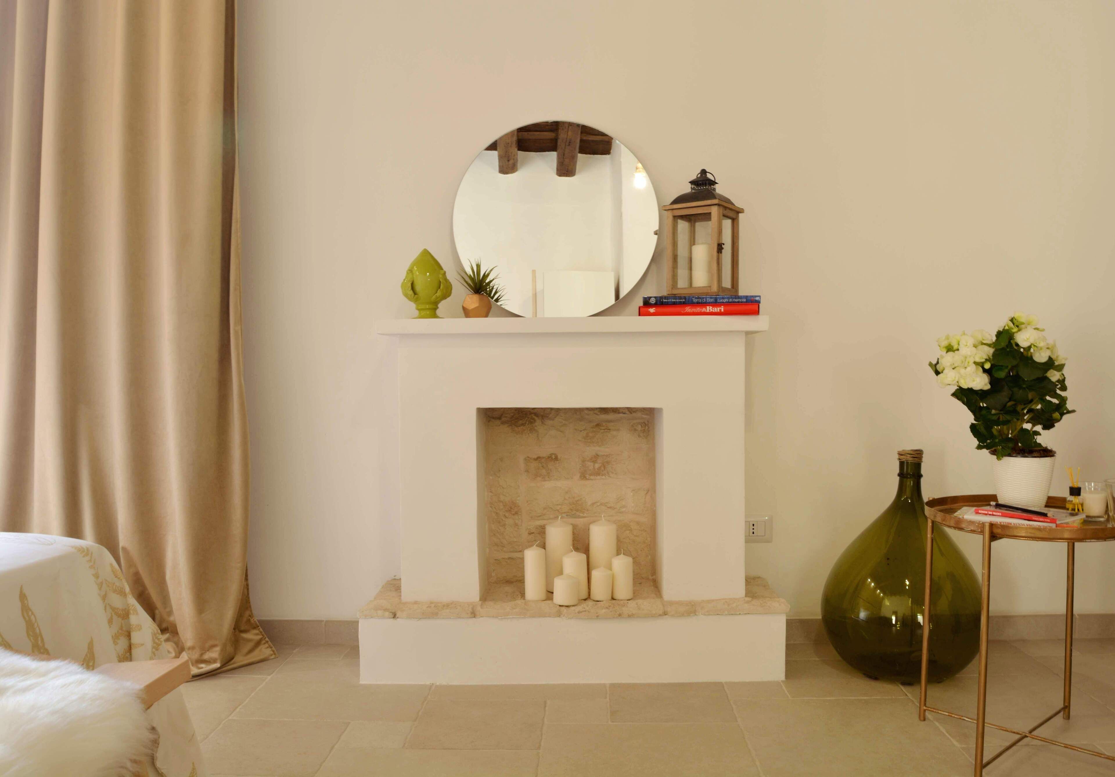 Una habitación con chimenea, un espejo, un estante pequeño con libros y un farol, y un gran jarrón verde con flores.