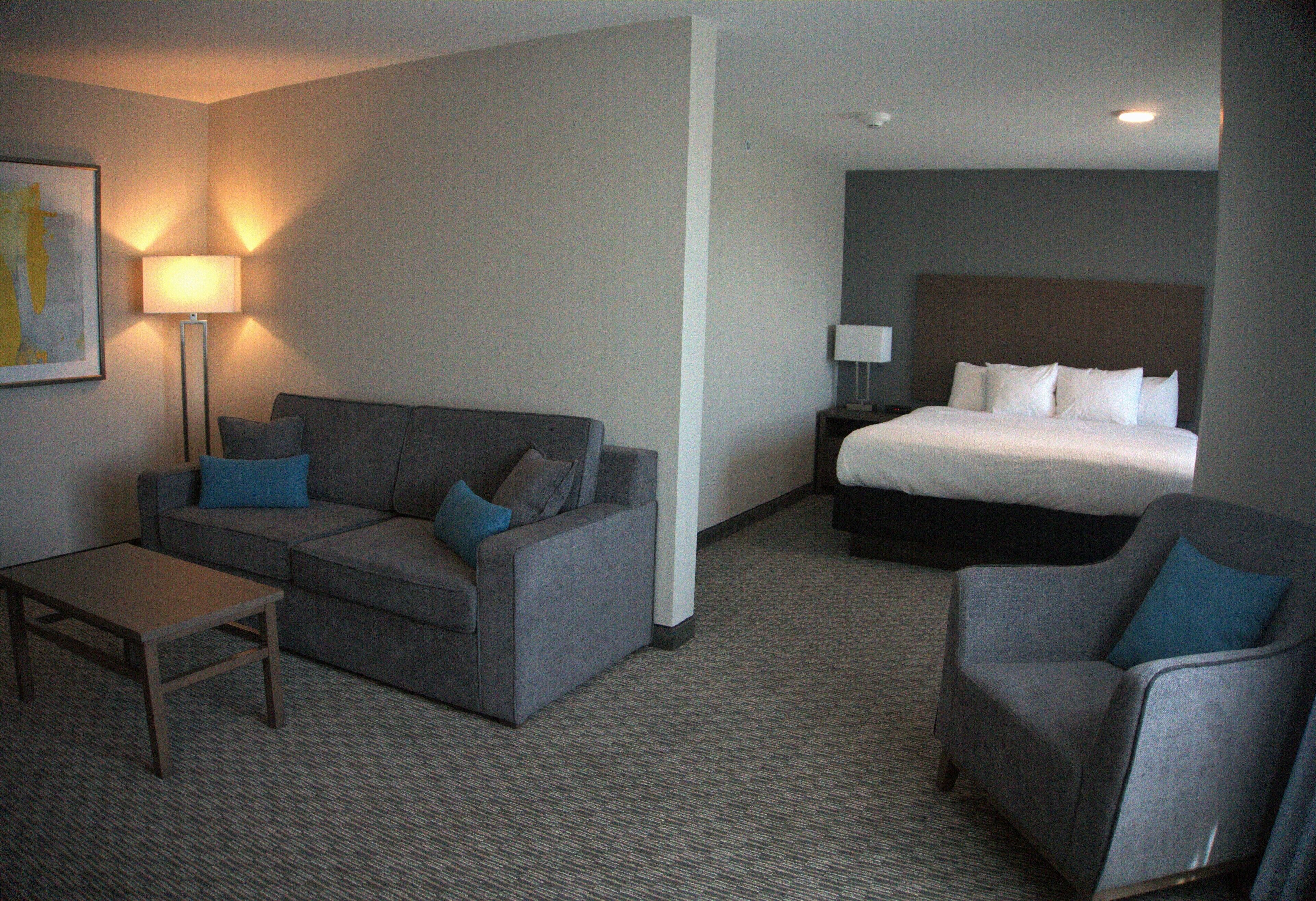 Une chambre d’hôtel avec un canapé gris, une petite table, un fauteuil gris, un lit avec une literie blanche et une table de chevet avec une lampe.