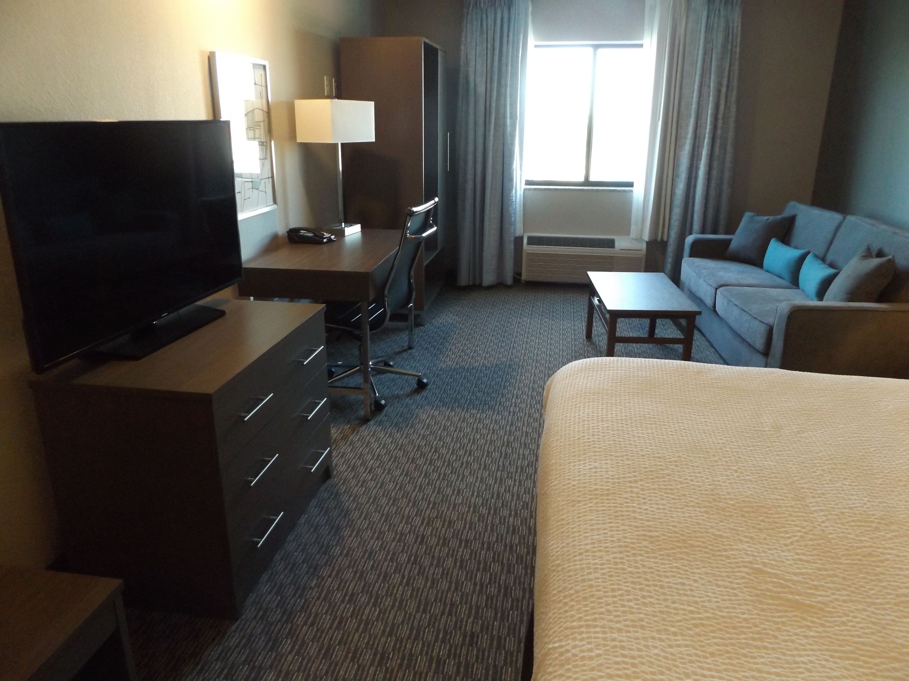 A hotel room with a bed, a desk with a chair, a television, a lamp, and a sofa.