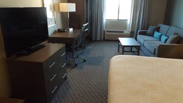 A hotel room with a bed, a desk with a chair, a television, a lamp, and a sofa.