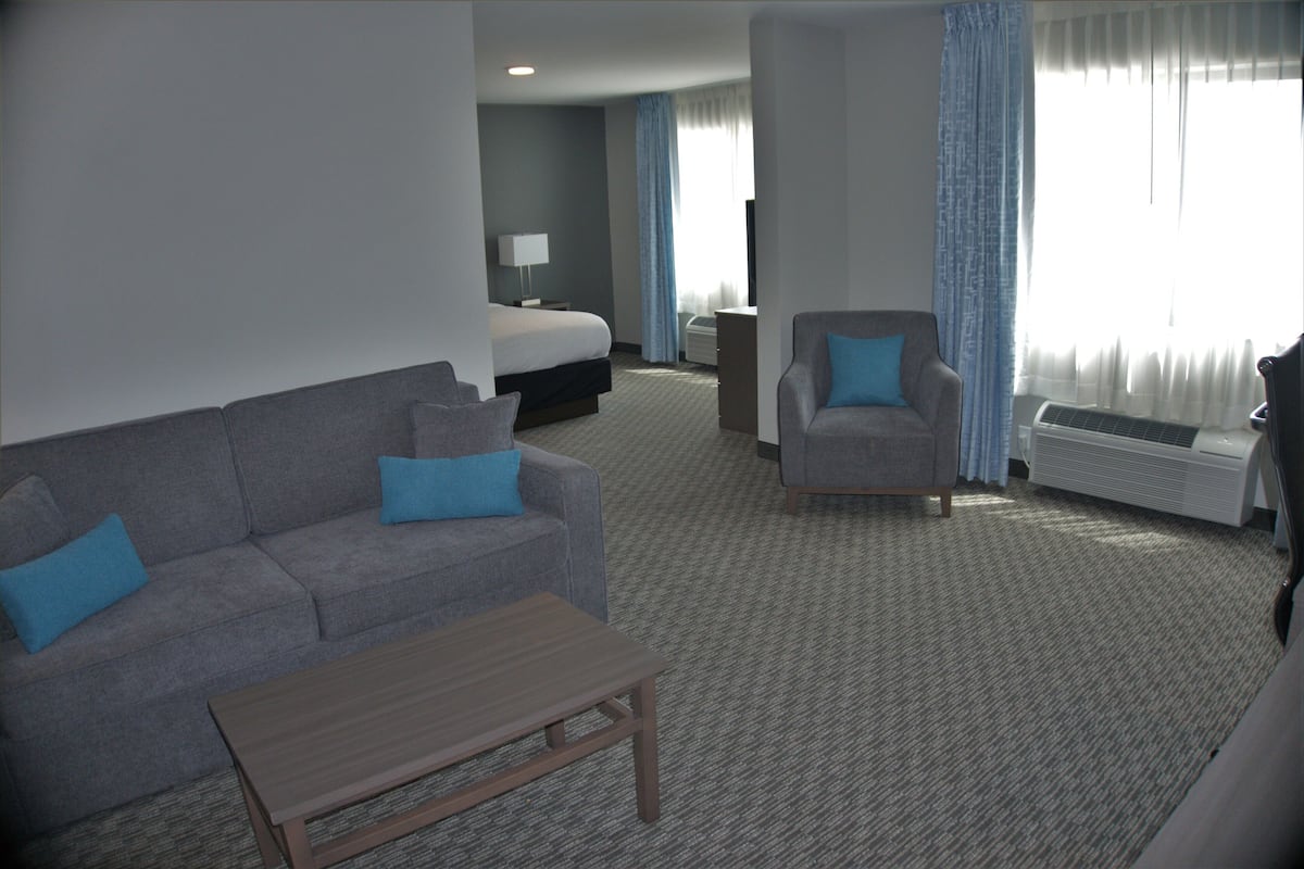 A hotel room with a grey sofa, a wooden coffee table, a grey armchair, a bed, a desk, and a window with curtains.
