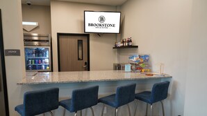 A bar area with a granite countertop, four blue bar stools, a refrigerator stocked with beverages, a television displaying the Brookstone logo, and shelves with snacks and bottled water. - Brookstone Fort Dodge (Fort Dodge)