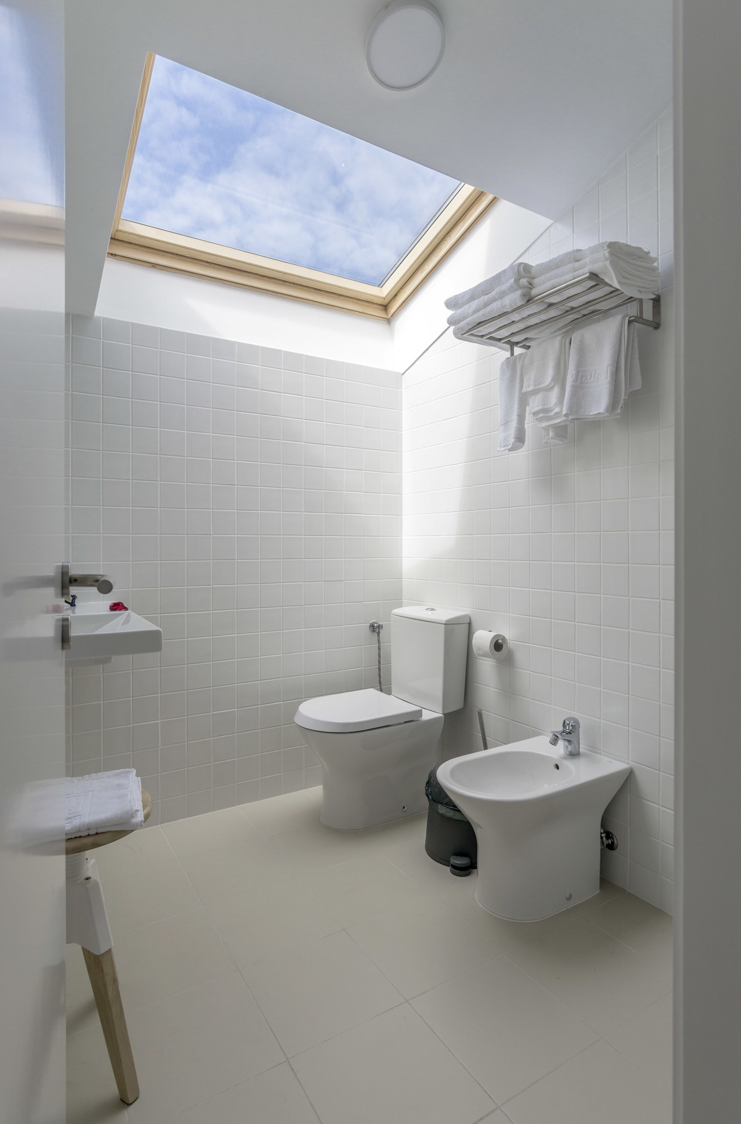 Une salle de bain moderne avec un puits de lumière, des carreaux blancs, des toilettes, un bidet et un porte-serviettes.