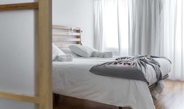 A neatly made bed with white linens and a gray blanket, a wooden headboard, and a window with sheer curtains.