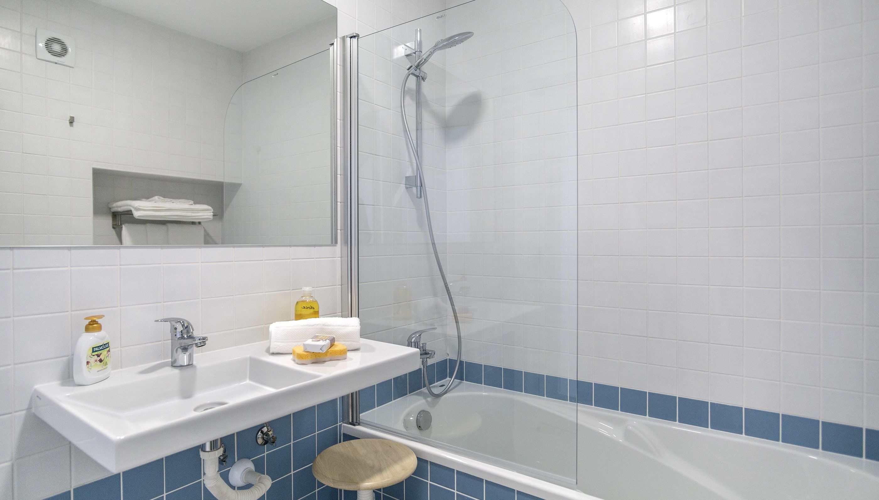 A bathroom with a white sink, mirror, and bathtub. There is a shelf with towels and toiletries.