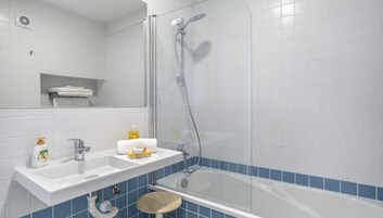 Un baño con lavamanos, espejo y bañera blancos. Hay un estante con toallas y artículos de aseo.