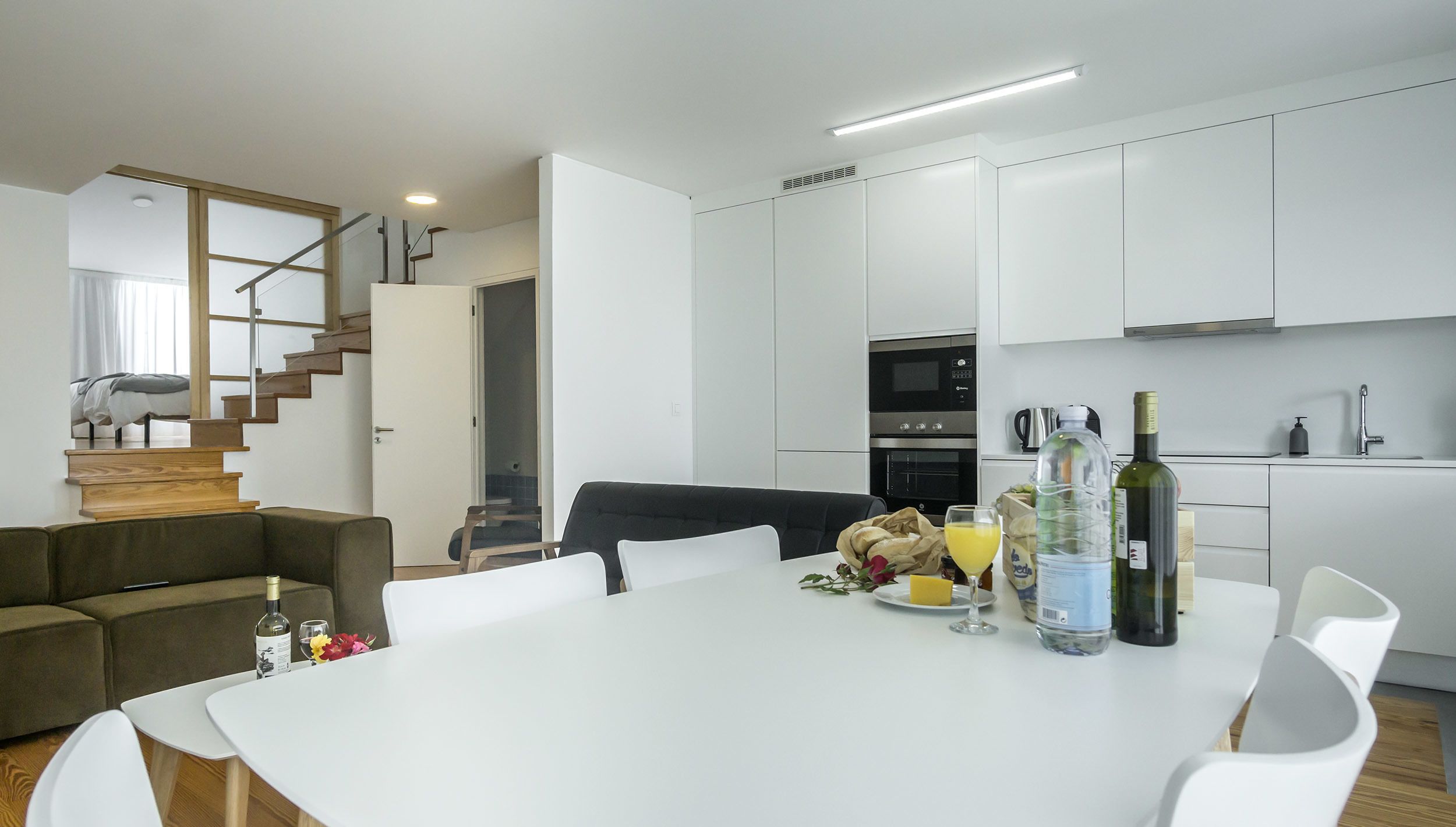 A modern kitchen with white cabinets, a dining area with a white table and chairs, and a staircase leading to another room.