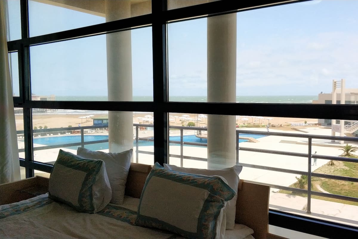 A hotel room with a large window overlooking a pool and beach area.