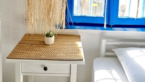 Une chambre avec un lit blanc, une petite table surmontée d’un set de table en fibres tressées, un luminaire suspendu et une fenêtre bleue avec des rideaux.