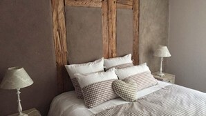 Una cama con colcha y almohadas a cuadros, flanqueada por dos mesitas de noche con lámparas, en una habitación con vigas de madera a la vista y una pared lisa.