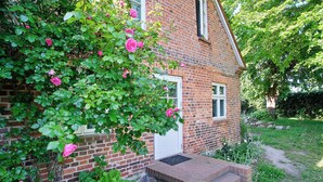 Ett hus byggt av tegel med en vit dörr och vita fönster, omgivet av en trädgård med grönt lövverk och rosa blommor.