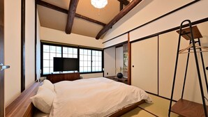 Une chambre de style japonais traditionnel, avec un lit, des poutres en bois, un revêtement de sol en tatami, une télévision et un placard à portes coulissantes.