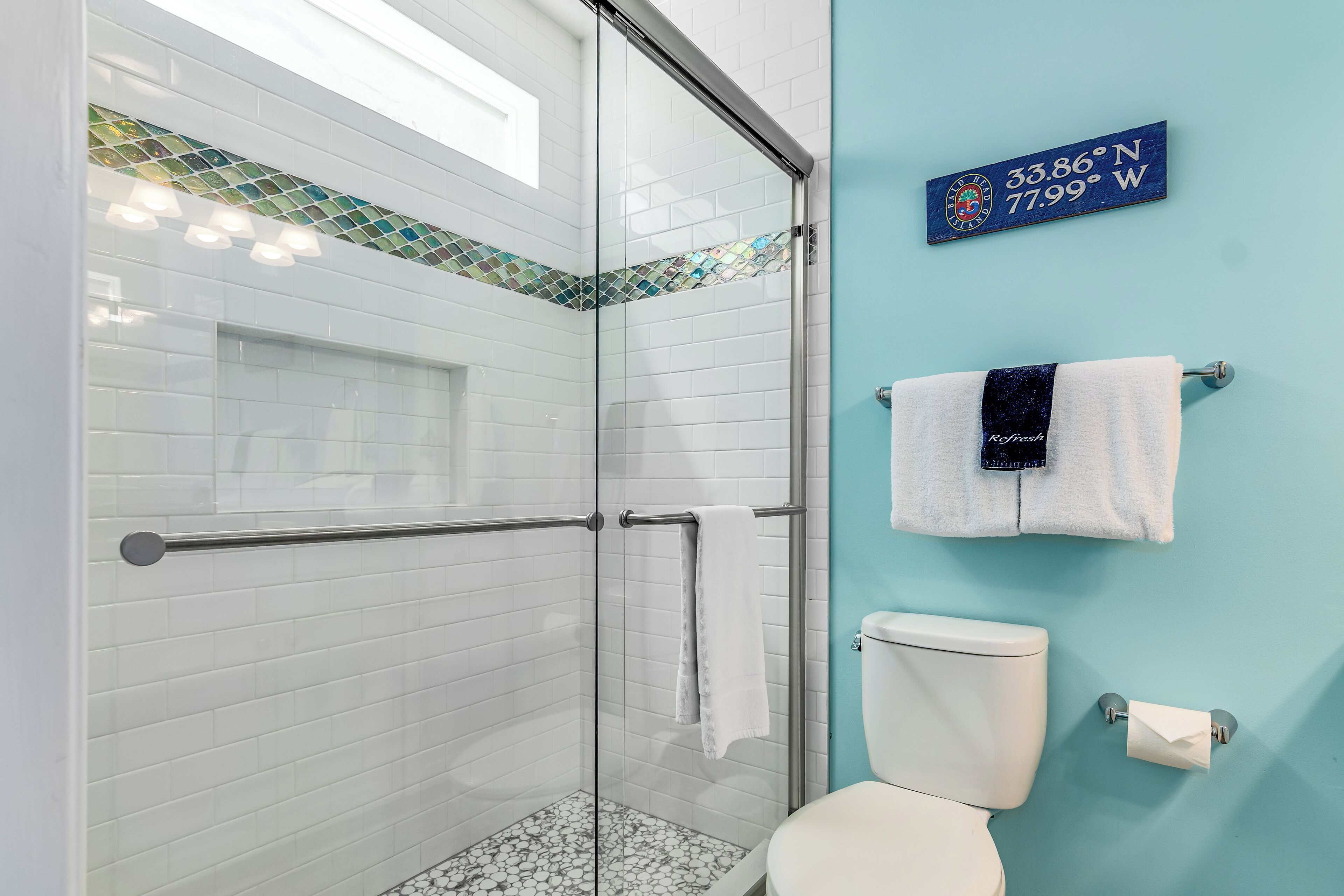 Ein Badezimmer mit Duschkabine, weißen Fliesen, einer blauen Akzentwand, Toilette und einem Handtuchhalter mit zwei Handtüchern.