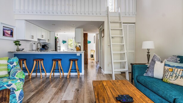 Amplia sala de estar con techos altos, una cocina con isla azul, pisos de madera y un sofá color verde azulado.