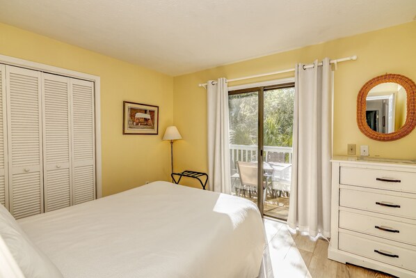 A bedroom with a bed, a dresser, a lamp, a mirror, and a sliding glass door to a balcony. - Condo with community pool and balcony with forest view, recently renovated (Bald Head Island)