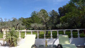 Un'area salotto all'aperto, luminosa, con due poltrone e un tavolo, circondata da un parapetto bianco e rigogliosa vegetazione.