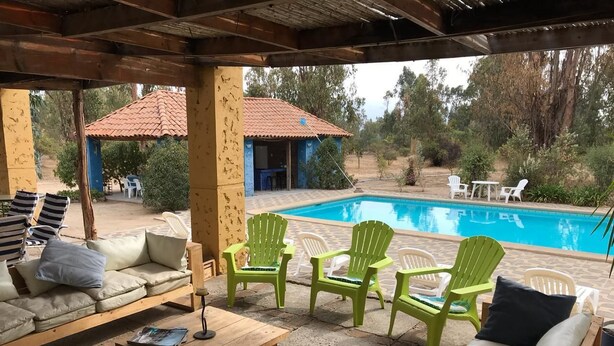 Un área de estar al aire libre con techo, piscina, sillas verdes y una mesa de madera.