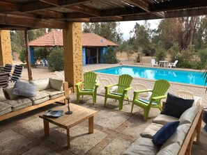 Un área de estar al aire libre con techo, piscina, sillas verdes y una mesa de madera.