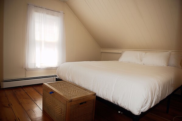 A bedroom with a large bed, wooden flooring, a window with curtains, and a wooden chest of drawers.