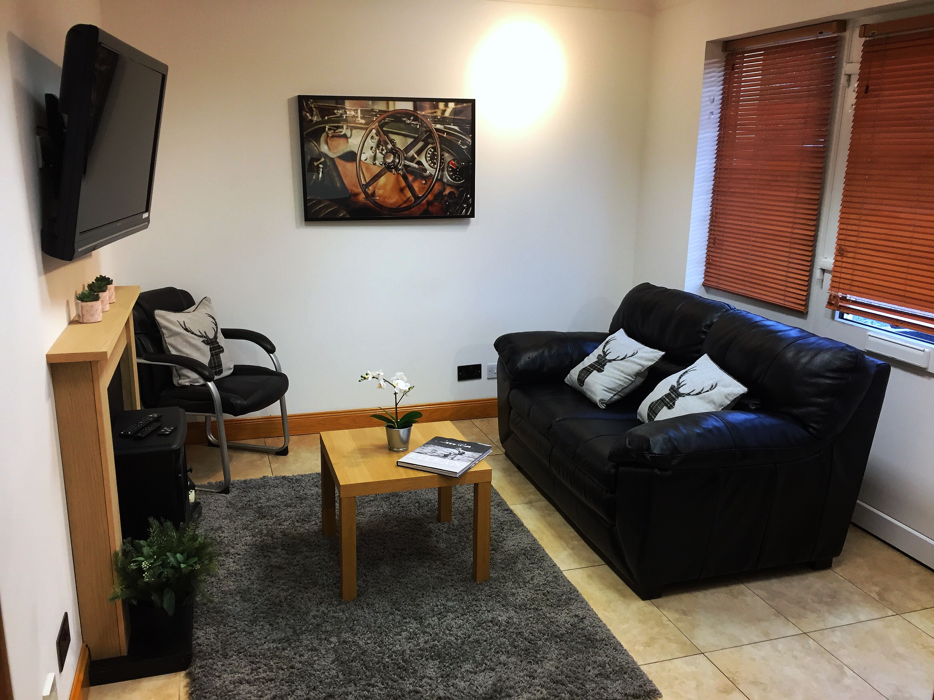 A living room with a black leather sofa, a wooden coffee table, a TV mounted on the wall, a painting, and a window with blinds.