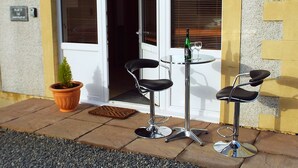 Un espace patio avec deux chaises et une table, une plante en pot et une bouteille de vin posée sur la table.