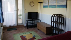 A living room with a television, a wooden bench, a chair, and a painting of a beach scene. - Spacious and cozy studio with breathtaking views (Saint-Mandrier-sur-Mer)