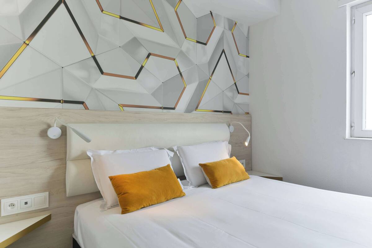 A modern bedroom with a geometric patterned wall, a wooden headboard, and two white beds with mustard-colored pillows.