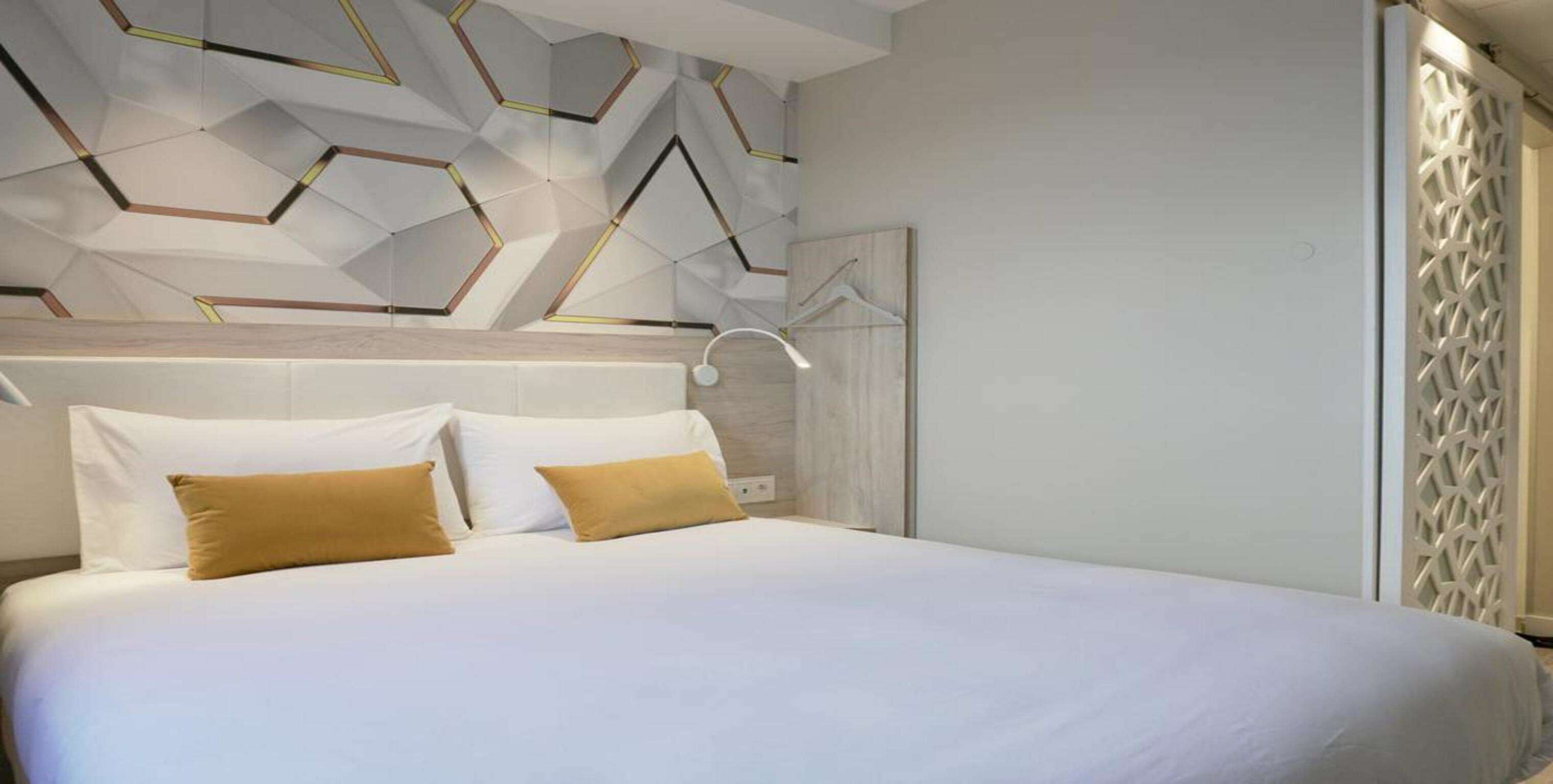 A modern hotel room with a geometric-patterned ceiling, a bed with white linens and two tan pillows, and a white headboard.