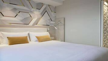A modern hotel room with a geometric-patterned ceiling, a bed with white linens and two tan pillows, and a white headboard.