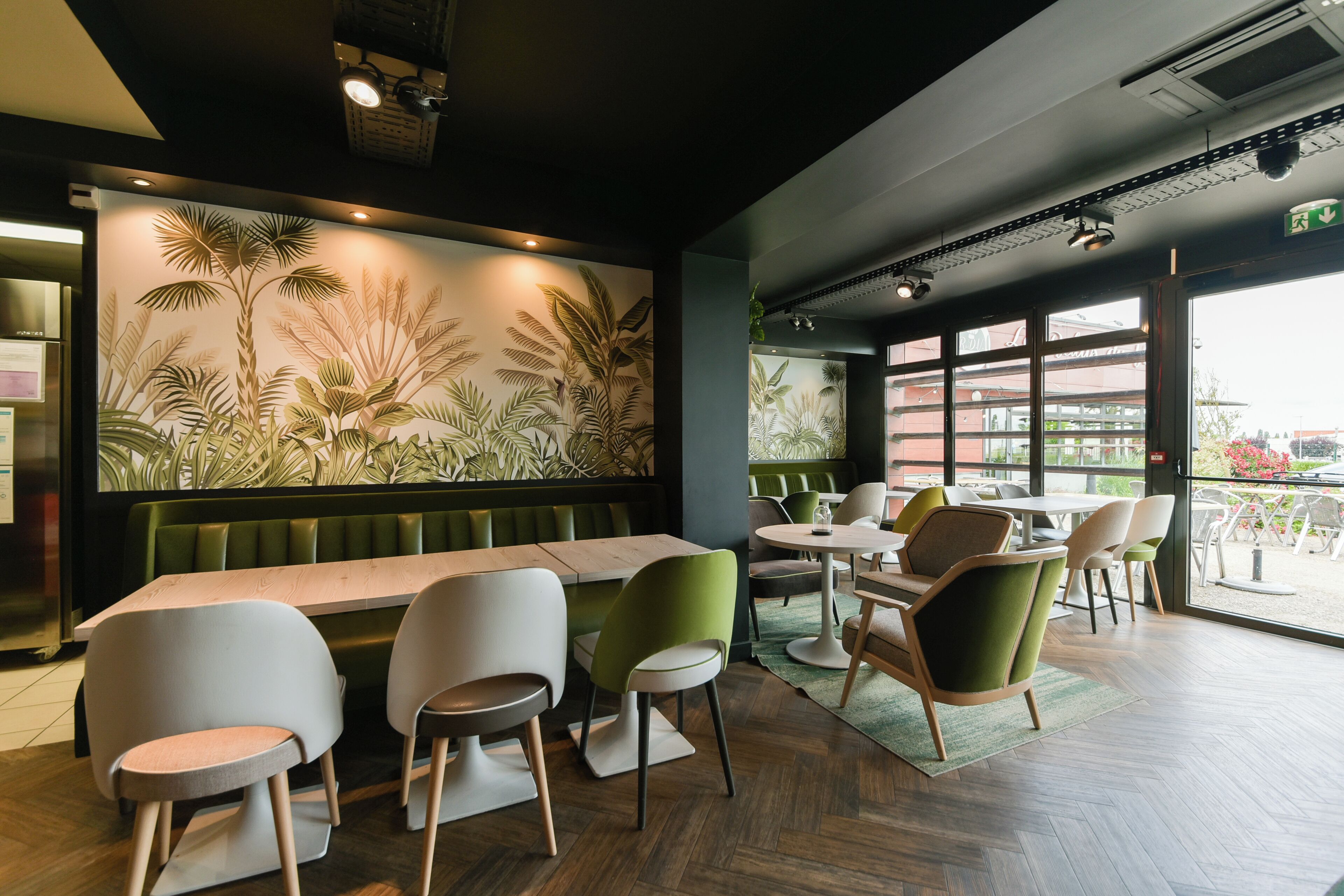 Interior de un restaurante con una larga mesa de madera, sillas verdes y blancas, y un gran mural en la pared con plantas tropicales.