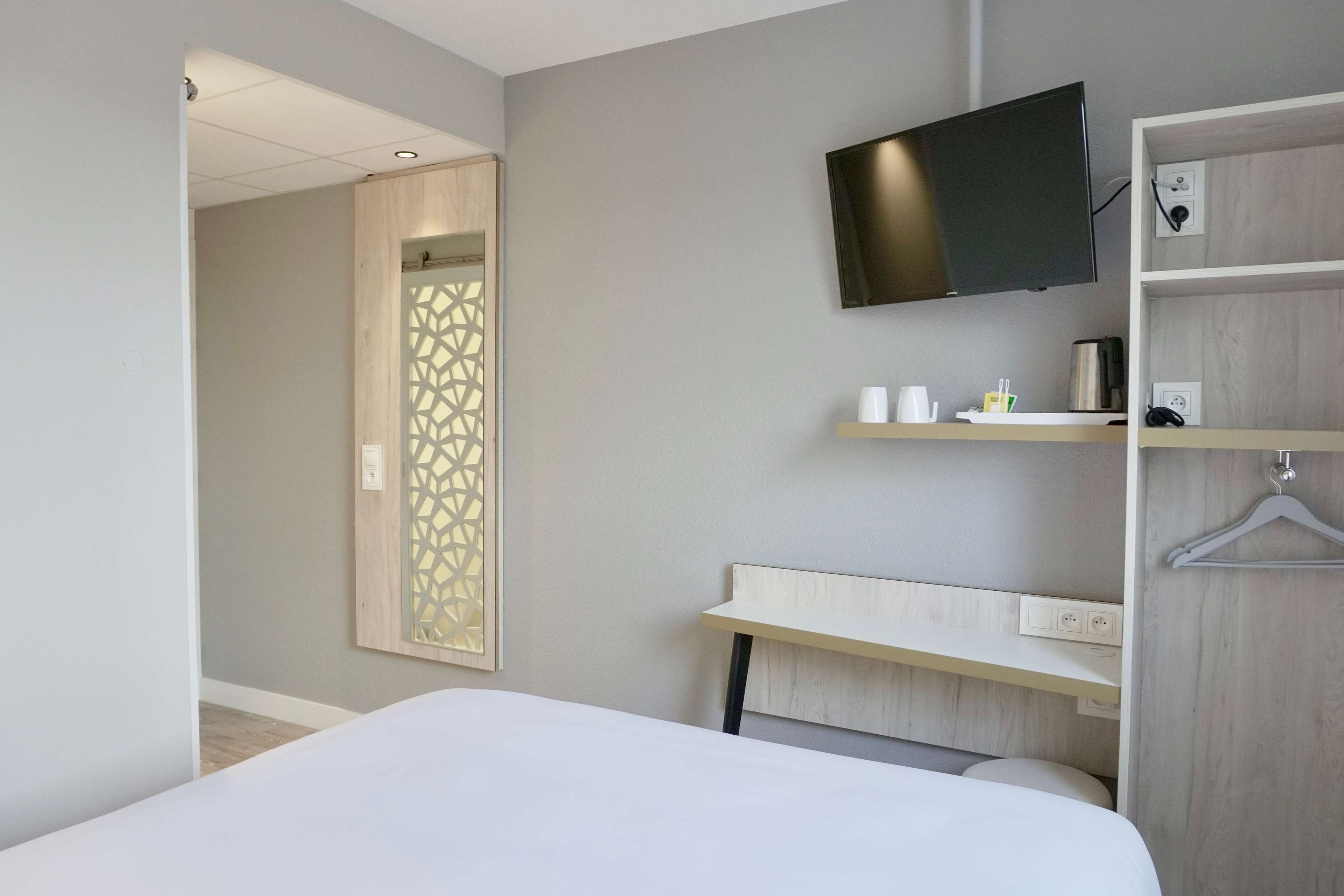 Una habitación de hotel moderna con una cama, un televisor montado en la pared, un escritorio con un portavasos y un espejo con diseño geométrico.