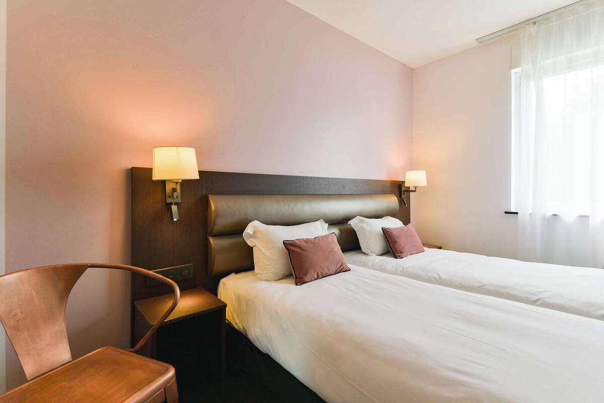 A hotel room with two beds, a wooden chair, and two wall-mounted lamps.