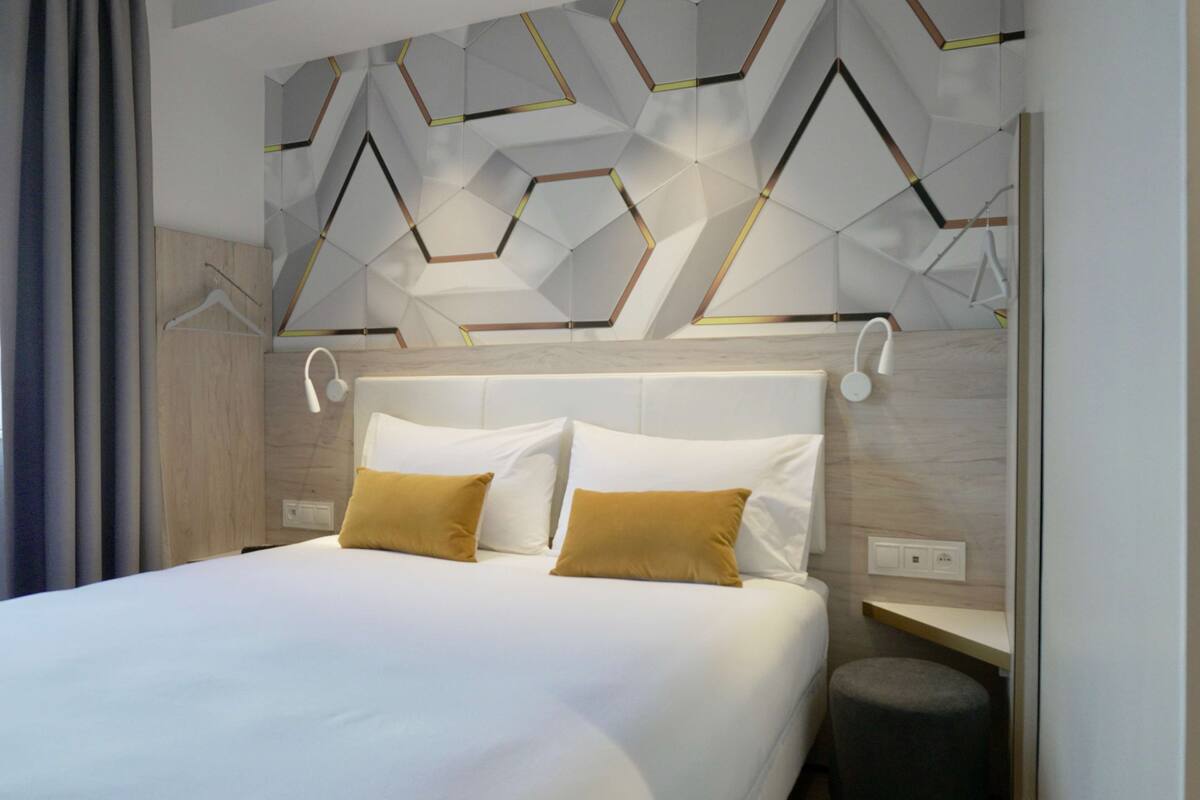 A modern hotel room with a geometric-patterned headboard, a neatly made bed with white and mustard pillows, and a small bedside table with a lamp.
