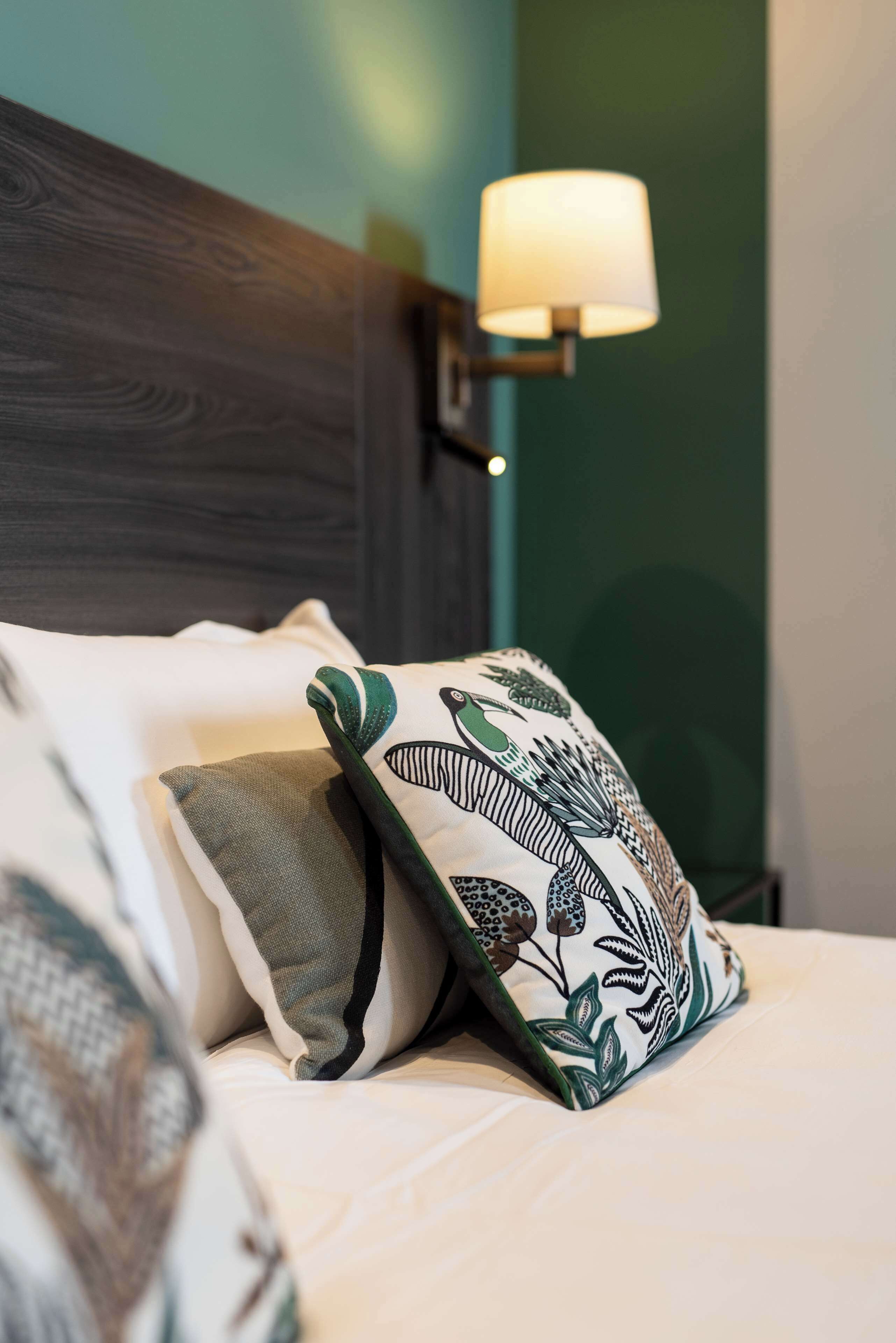 Un letto rifatto con cura, con una testiera in legno scuro, una parete verde e una lampada fissata al muro.