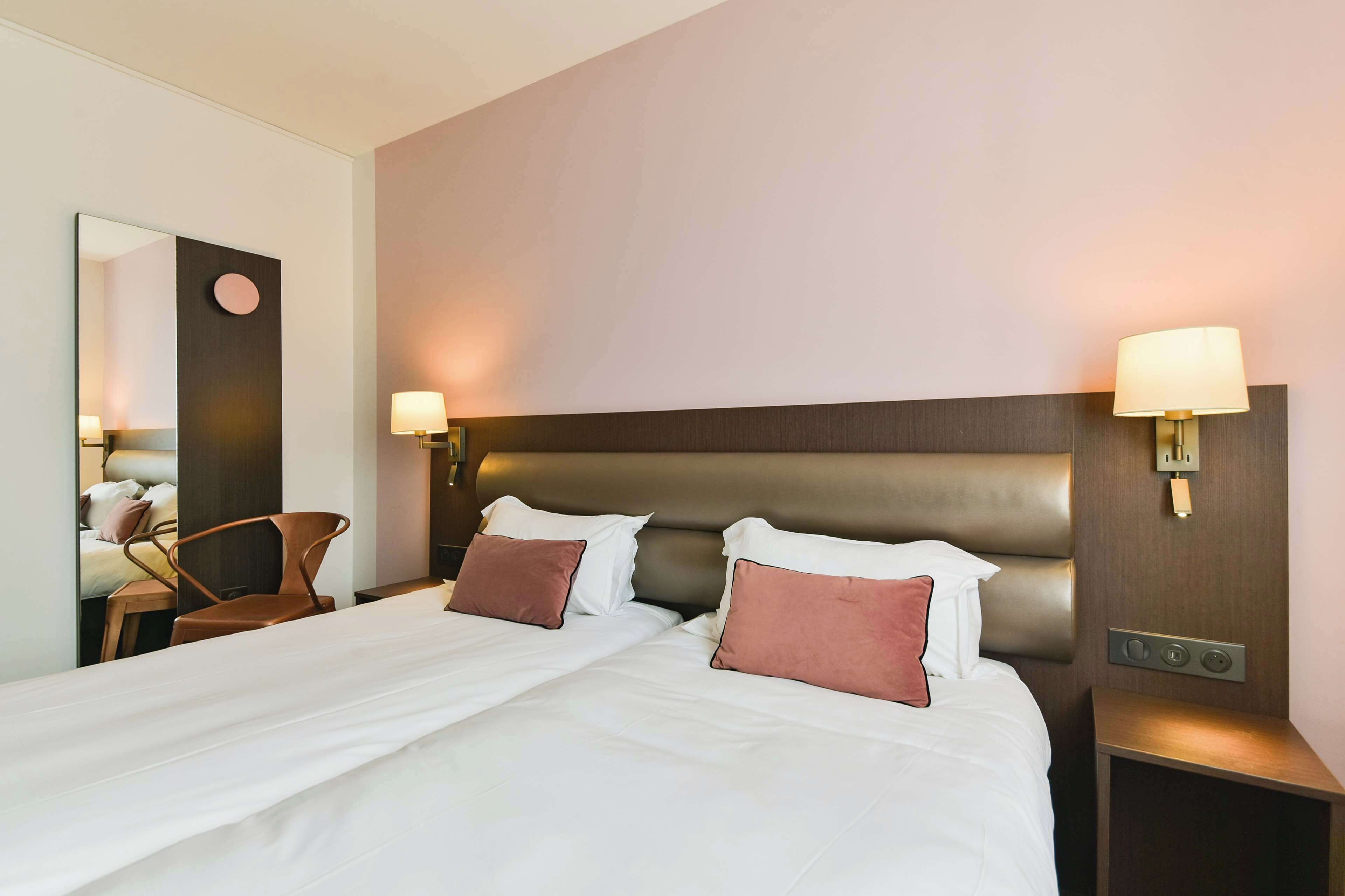 A neatly made bed with white and brown bedding, a wooden nightstand with a lamp, and a wall-mounted light fixture.