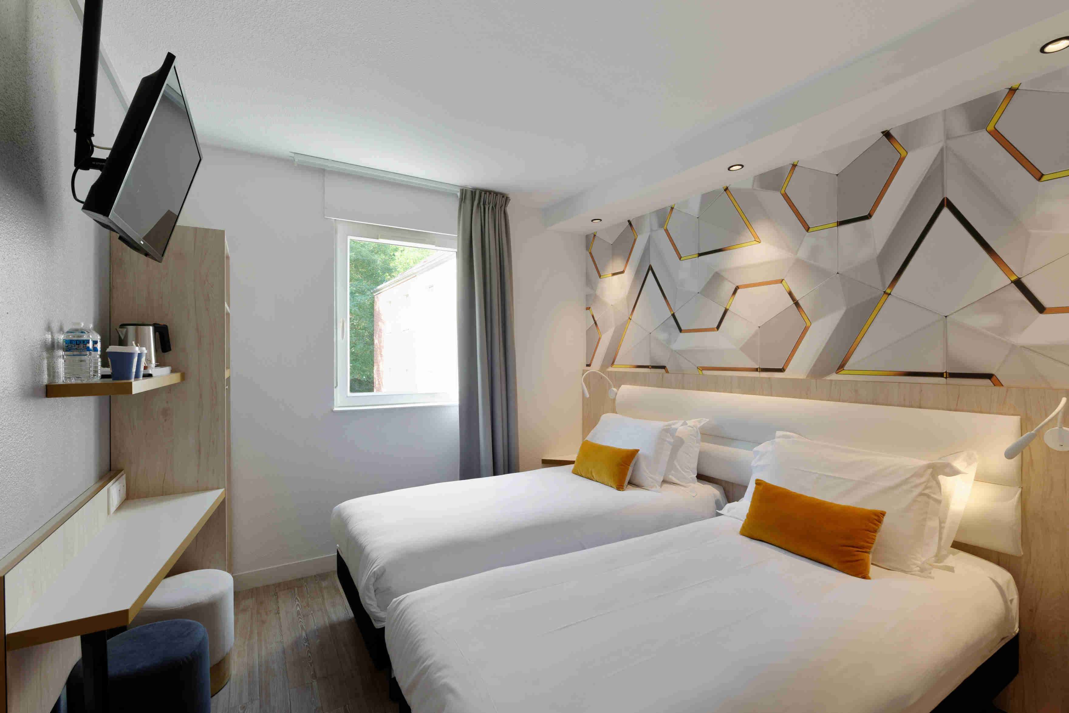Una habitación de hotel moderna con dos camas, un escritorio, un televisor y un diseño geométrico en la pared.