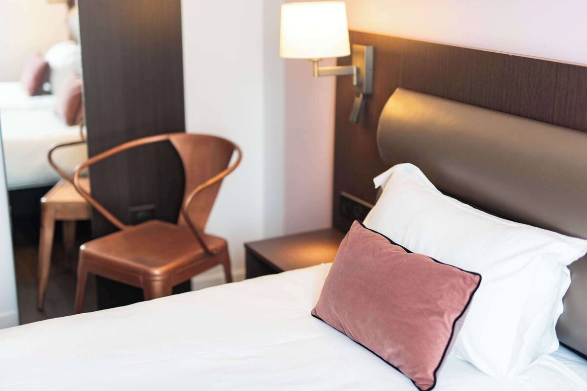 A hotel room with a bed, a chair, a nightstand, and a wall-mounted lamp.