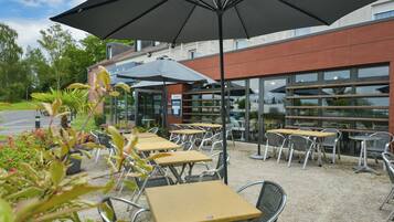 Een terras met tafels en stoelen, een grote parasol en een gebouw met grote ramen op de achtergrond.