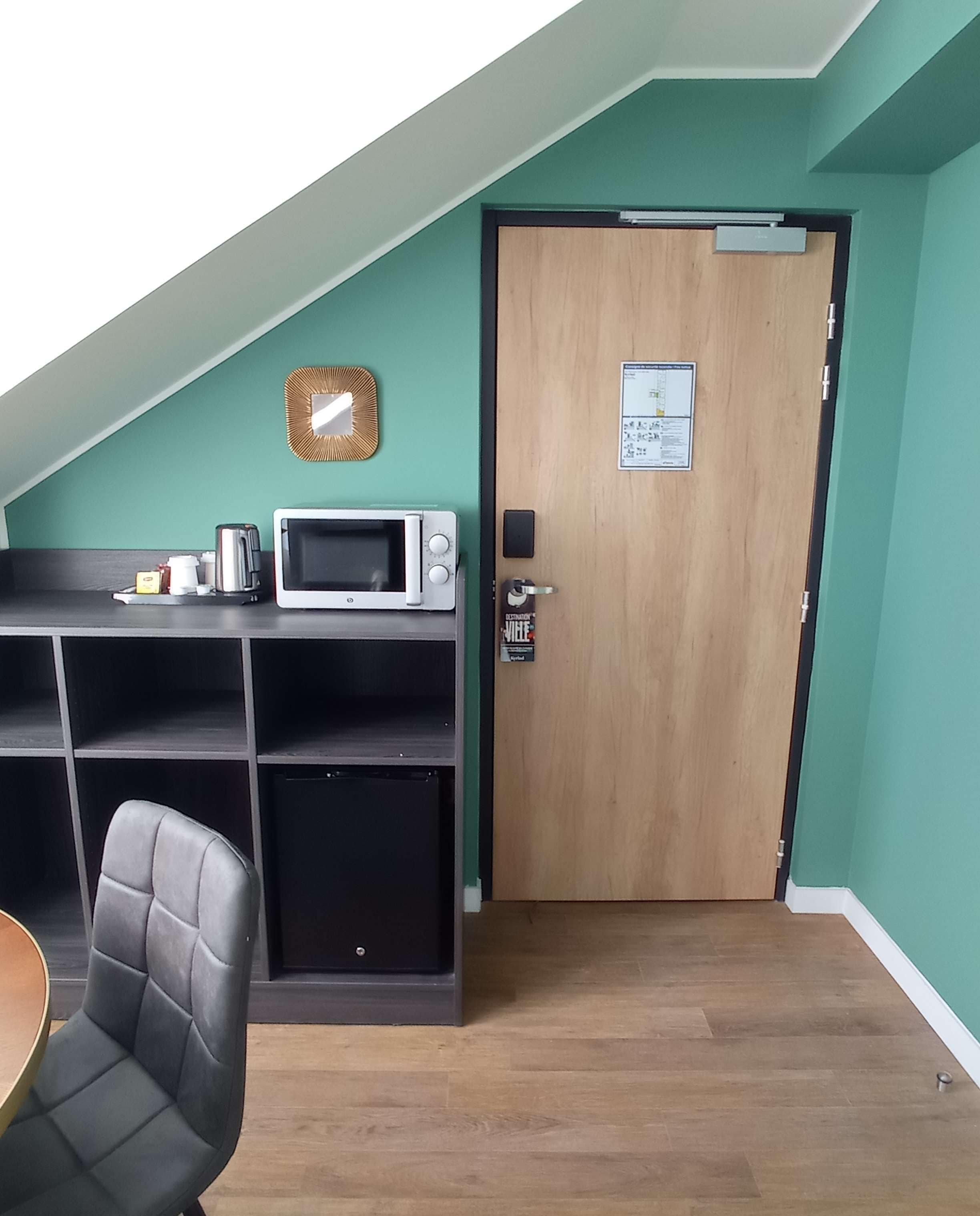Ein Zimmer mit Holztür, einer Mikrowelle, einem Regal mit verschiedenen Gegenständen und einem Stuhl.
