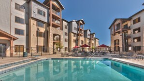 A swimming pool surrounded by multi-story buildings with balconies, outdoor seating, and a clear sky.