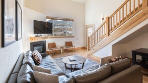 A modern living room with a sofa, a coffee table, and a TV. There is a staircase with wooden railings and a view of the outside.