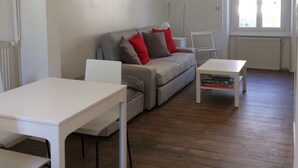 A modern living room with a grey sofa, white chairs, a white table, and a window with blinds. - Apartment T2 - LOW SABLONS (Saint-Malo)