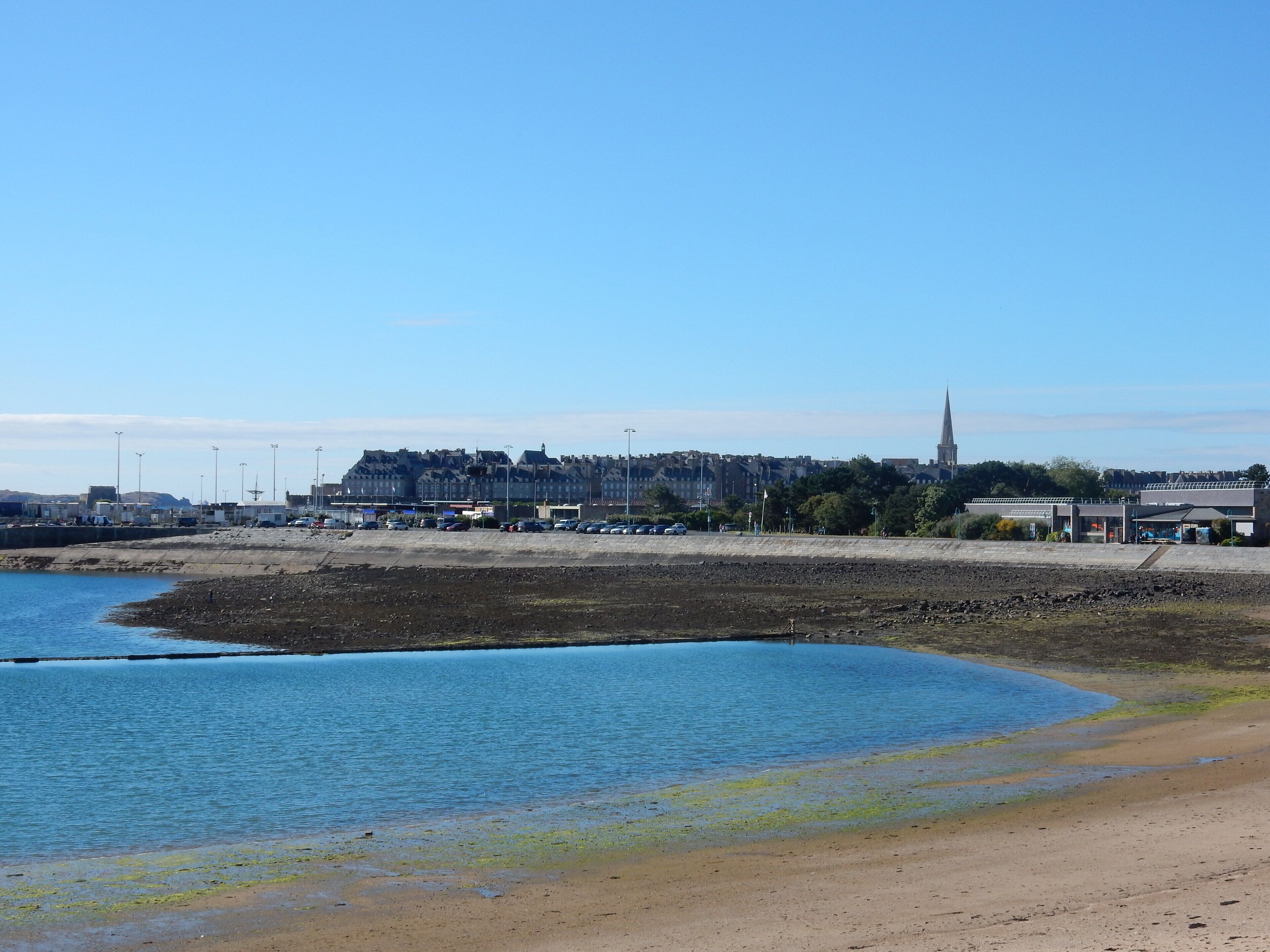 A coastal area with a sandy beach, a calm blue body of water, and a cityscape in the background.
