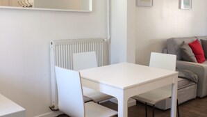 A modern, minimalist living room with a white dining table and chairs, a grey sofa with red cushions, and a white radiator. - Apartment T2 - LOW SABLONS (Saint-Malo)