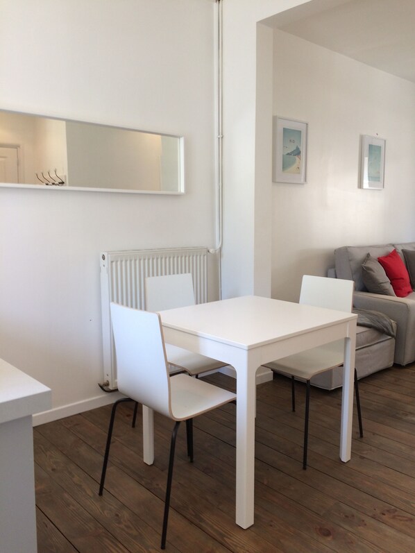 A modern, minimalist living room with a white dining table and chairs, a grey sofa with red cushions, and a white radiator.